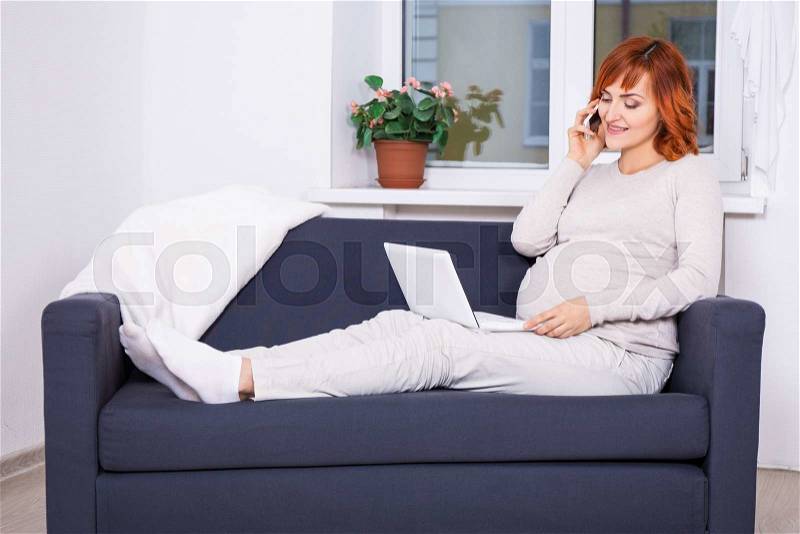 Young beautiful pregnant woman using laptop and talking by phone in living room, stock photo