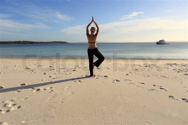Woman balanced in a yoga pose standing barefoot on one leg with her hands joined above her head at the beach in late afternoon sunlight, drawing long shadows on the sand. Asana vrksasana, stock photo