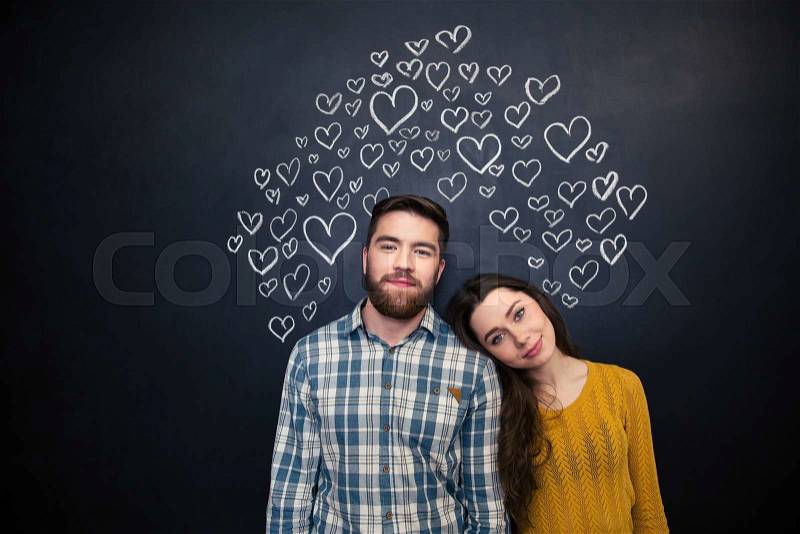 Beautiful tender young couple standing over black board with drawn hearts behind them, stock photo