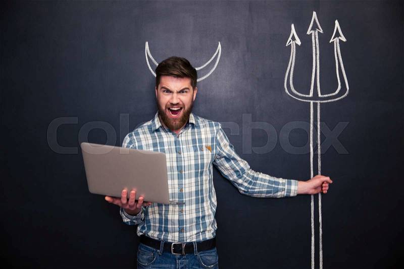 Angry bearded man using laptop and holding trident drawn on blackboard background, stock photo