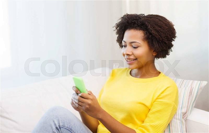 People, technology and leisure concept - happy african american young woman sitting on sofa with smartphone at home, stock photo