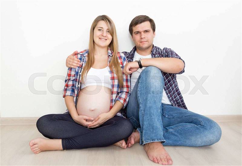 Cute pregnant couple in jeans and shirts sitting on floor at empty room, stock photo