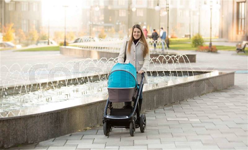 Happy young mother with baby stroller walking on street near fountain, stock photo