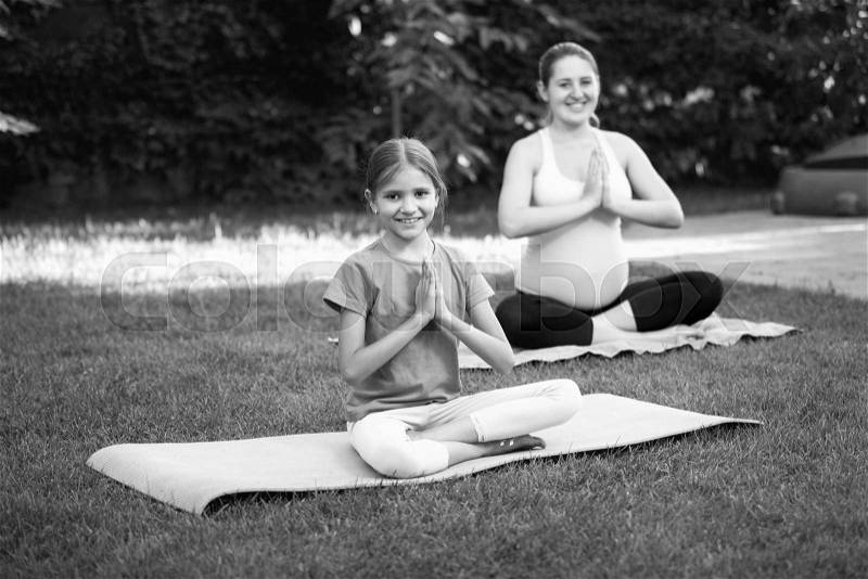 Black and white photo of pregnant mother with daughter practicing yoga on grass at park, stock photo