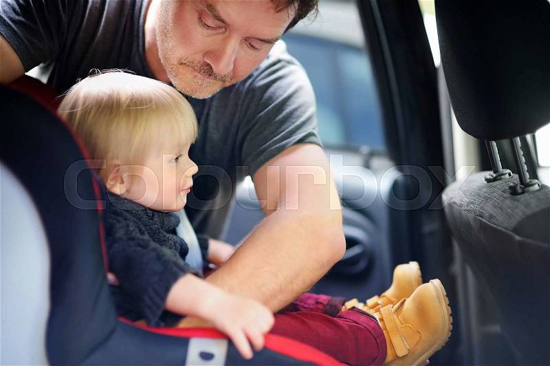 Middle age father helps his toddler son to fasten belt on car seat, focus on father, stock photo