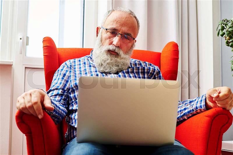 Sad senior man sitting on the chair and looking at laptop, stock photo
