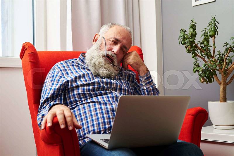 Bearded senior man sleeping with laptop on the red chair at home, stock photo