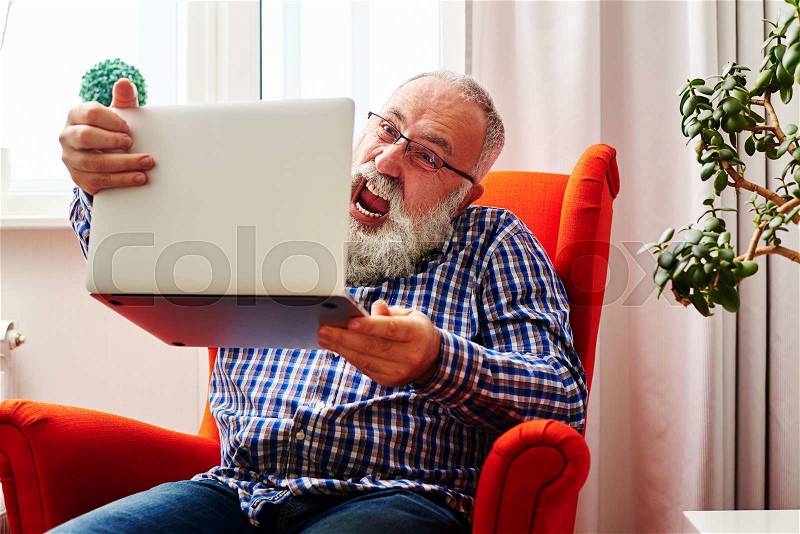 Mad senior man sitting on the chair and screaming at laptop at home, stock photo