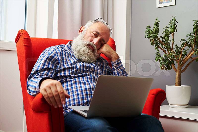 Tired senior man sleeping with laptop on the chair at home, stock photo