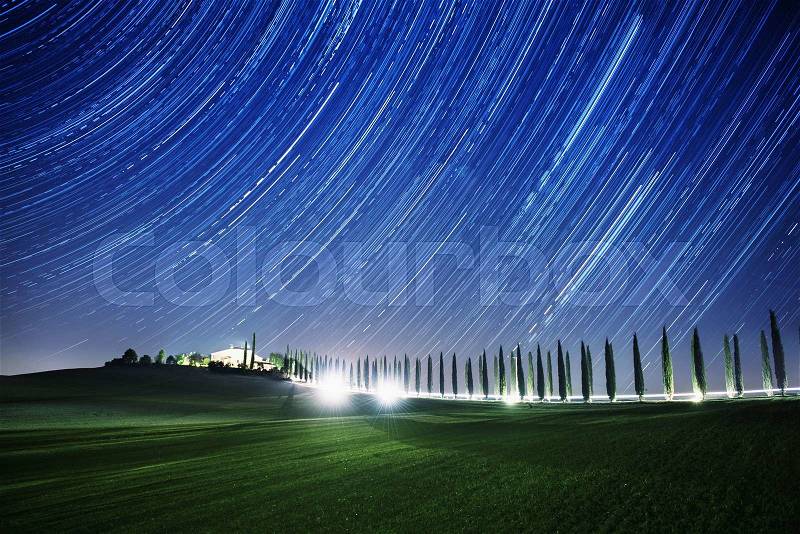 Beautiful Tuscany night landscape with star trails on the sky, cypresses and shining road in green meadow. Natural outdoor amazing fantasy background, stock photo