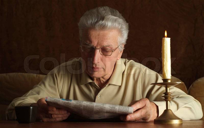 Portrait of a mature man reading newspaper with candle, stock photo