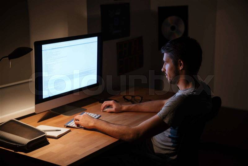 Handsome young man sitting in dark room and using computer, stock photo