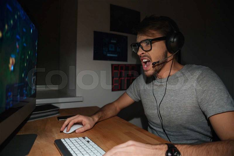 Excited handsome young gamer in glasses and earphones playing computer game at home, stock photo