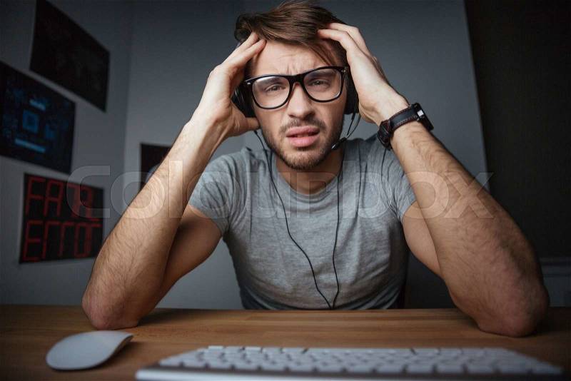 Frowning young man in earphones and glasses sitting with hands on head in front of computer, stock photo