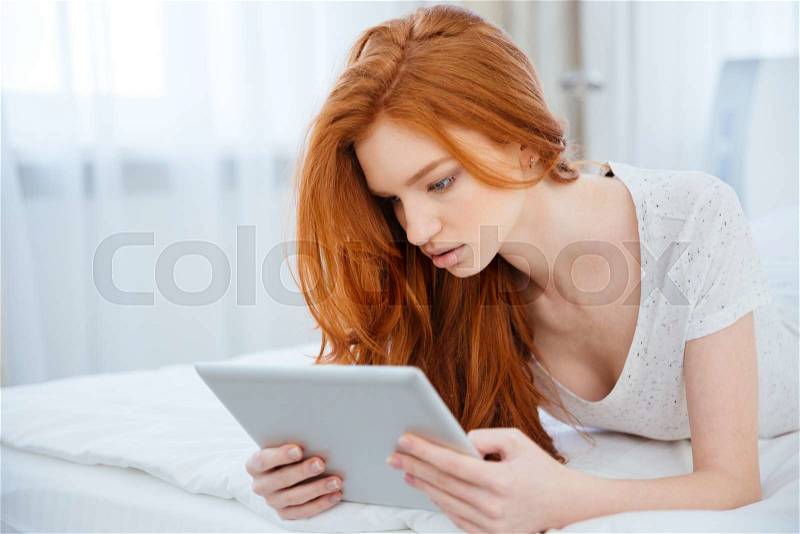 Beautiful redhead woman using tablet computer on the bed at home, stock photo