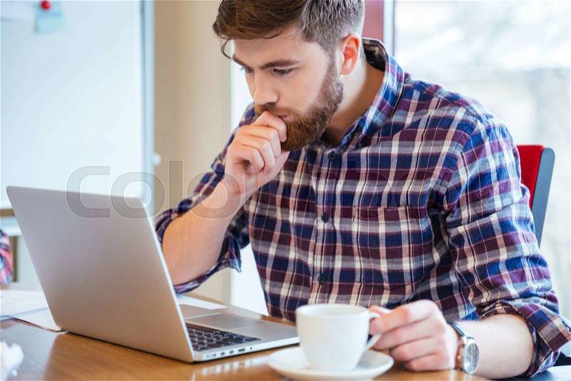 Serious focused bearded young man in checkered shirt sitting at the table and using laptop, stock photo