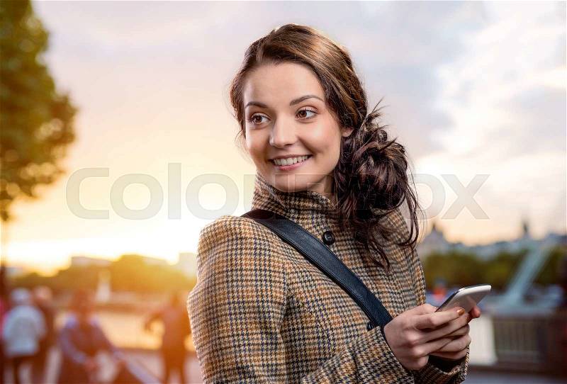 Young woman in checked brown winter coat with smart phone outside in sunny park, stock photo