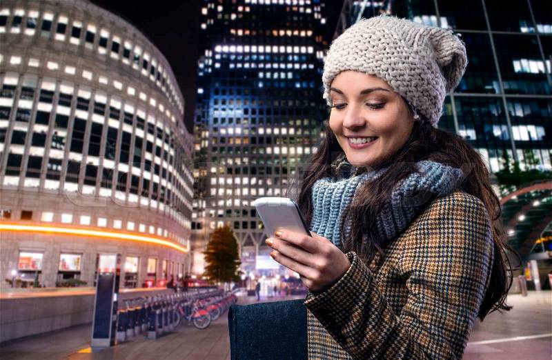 Young woman in checked brown winter coat with smart phone outside in illuminated night city, stock photo