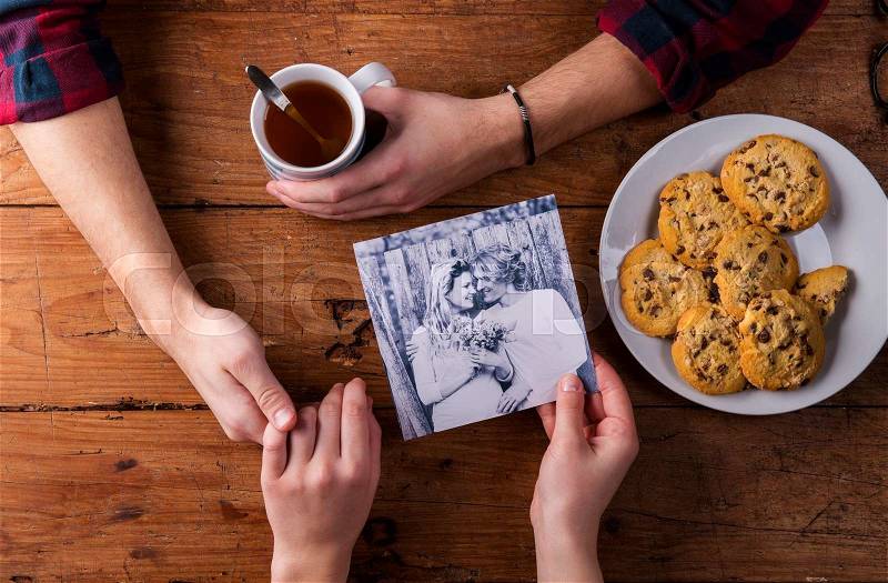 Unrecognizable man holding womans hand. Looking at their black and white photo. Couple in love. Cookies and tea. Valentines day composition. Studio shot on brown wooden background, stock photo