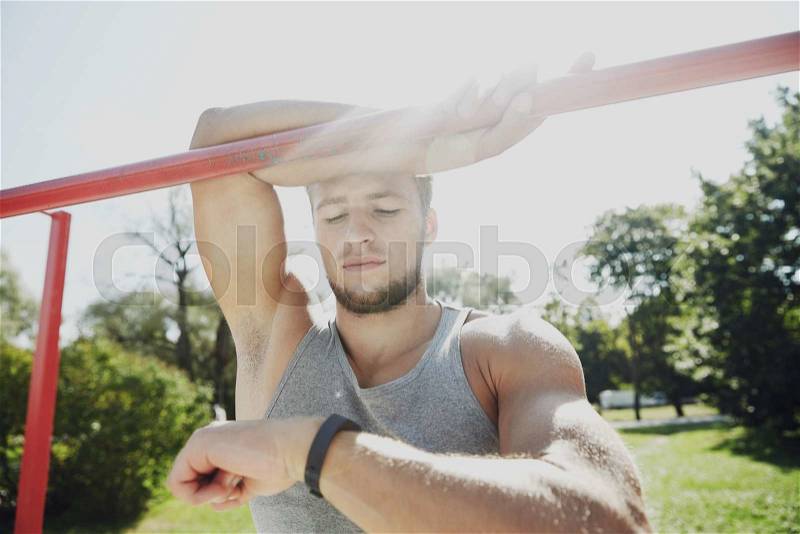 Fitness, sport, training and lifestyle concept - happy young man looking at heart-rate watch bracelet and exercising on horizontal bar in summer park, stock photo