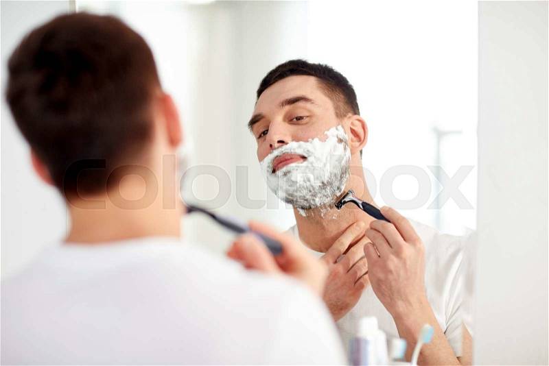 Beauty, hygiene, shaving, grooming and people concept - young man looking to mirror and shaving beard with manual razor blade at home bathroom, stock photo