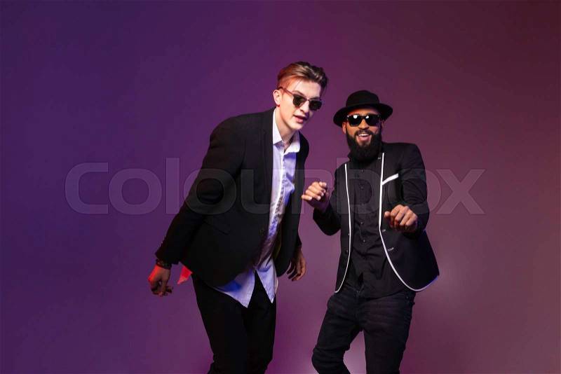 Two attractive cheerful young men in sunglasses dancing over purple background, stock photo