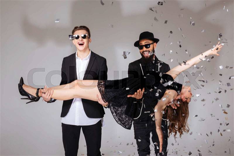 Two smiling young men standing and holding attractive relaxed young woman over white background, stock photo