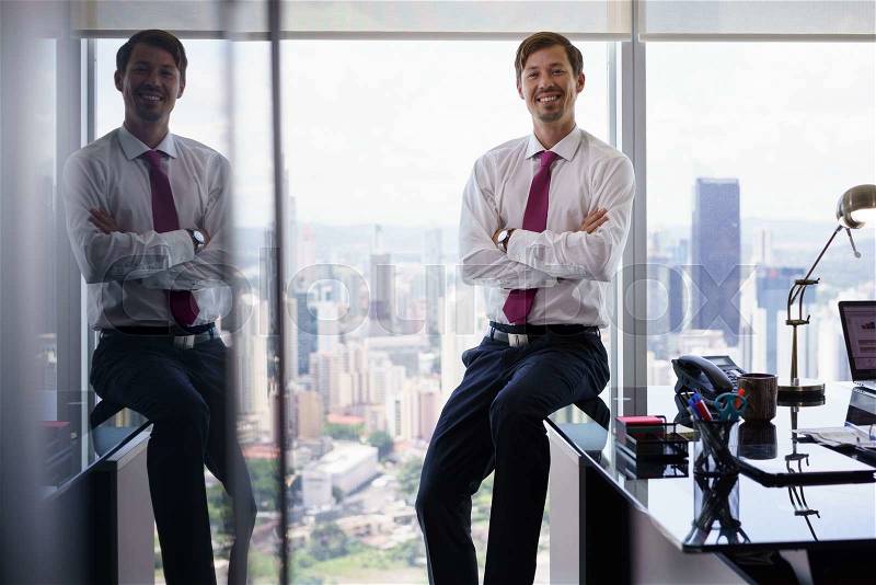 Adult businessman sitting in modern office with beautiful sight of the city. The man looks at the camera and smiles with arms crossed, stock photo