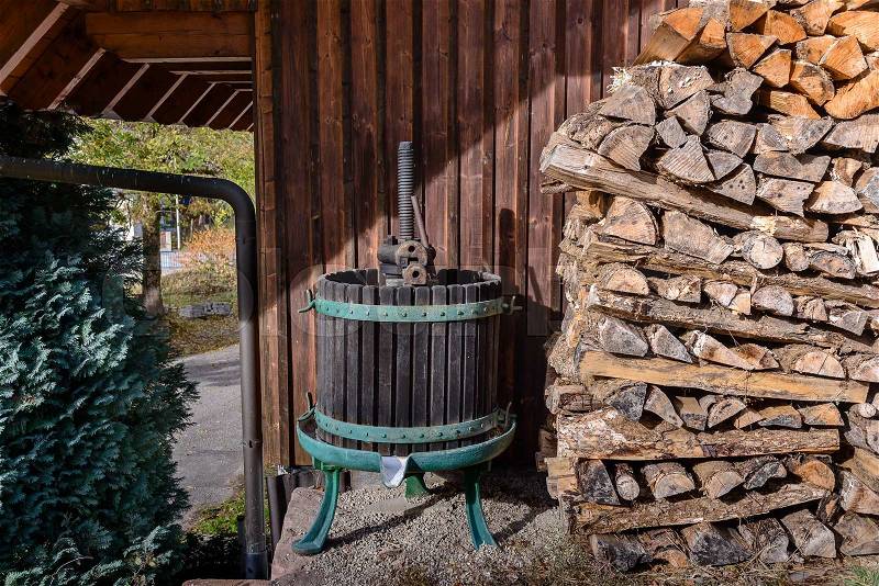 Old fashioned wine press next to pile of cut logs next to wall near garage with tree and gutter, stock photo