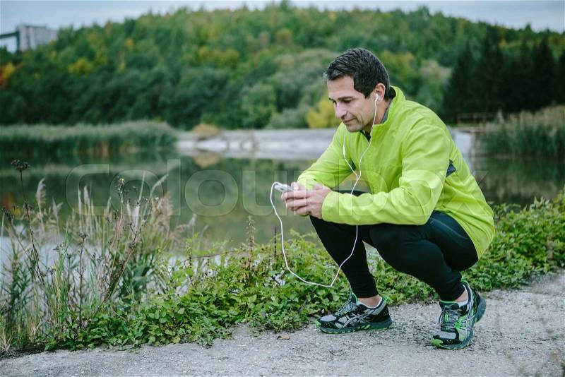 Runner with smart phone and earphones, in yellow neon jacket, on the path at the lake resting against green nature, stock photo