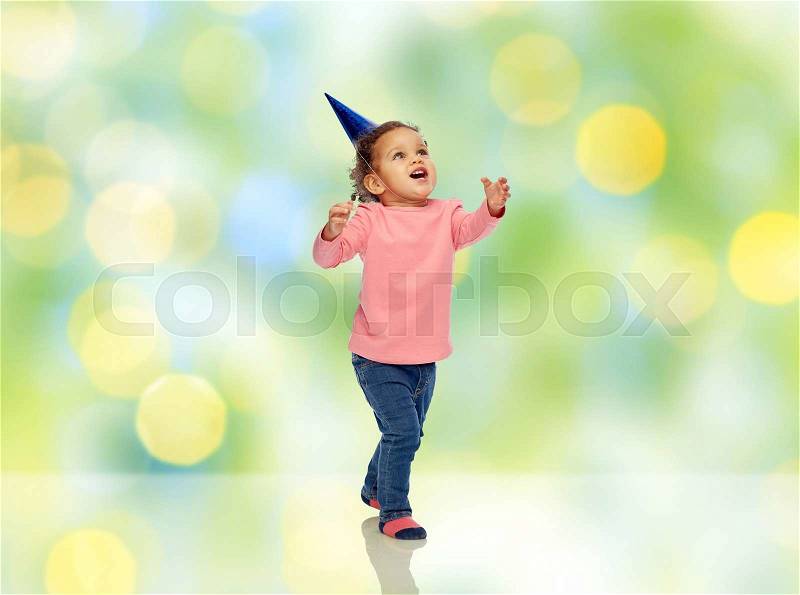 Childhood, fashion, birthday, holidays and people concept - happy smiling african american little baby girl with birthday party hat catching something over green summer holidays lights background, stock photo