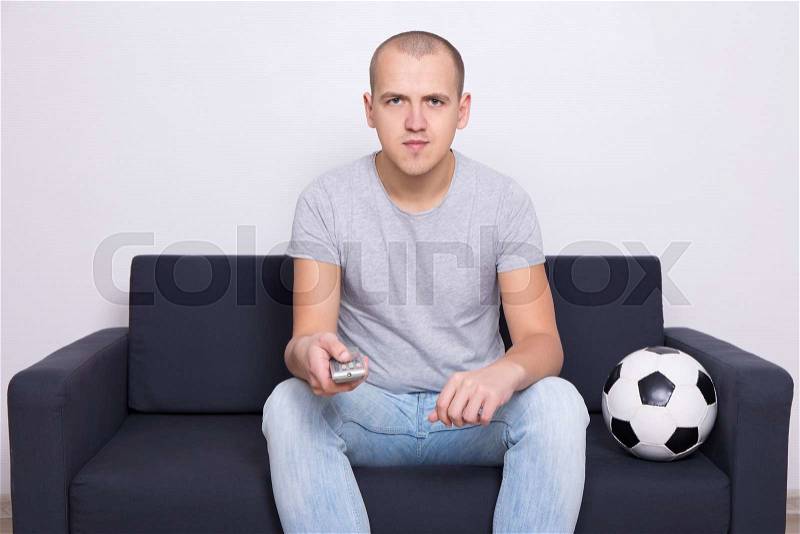 Young man sitting on sofa with soccer ball and watching tv at home, stock photo