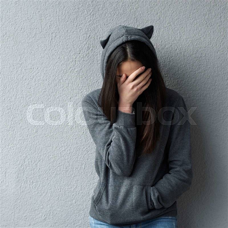 Sad woman in grey hoodie with cat\'s ears covering face with hand. Wall background, stock photo
