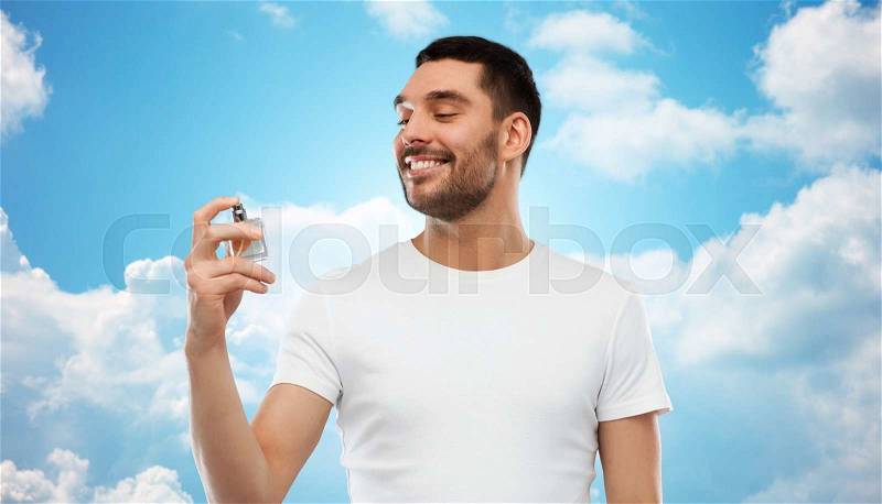 Perfumery, beauty and people concept - happy smiling young man with male perfume over blue sky and clouds background, stock photo