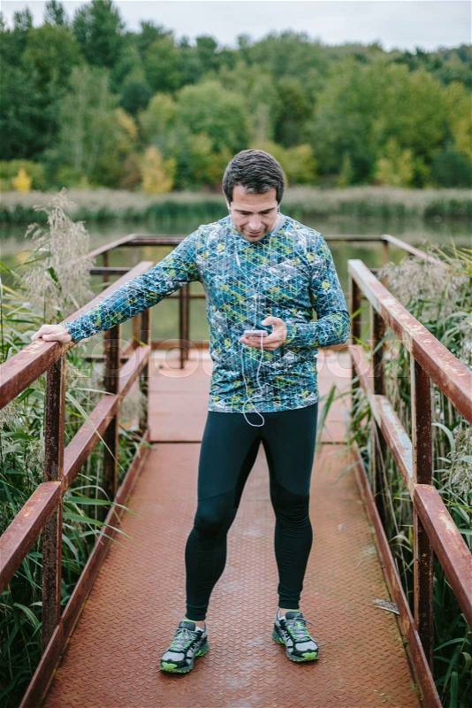 Runner on an old rusty metal bridge holding smart phone and earphones, listening music, against green nature, stock photo