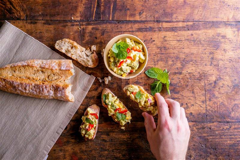 Spread made of avocado and other ingredients in a bowl and on slices of bruschetta. Hand of man taking it, eating. Studio shot on old wooden table background , stock photo