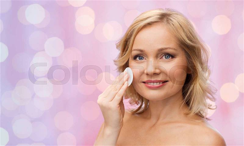 Beauty, people and skincare concept - young woman cleaning face and removing make up with cotton pad over pink lights background, stock photo