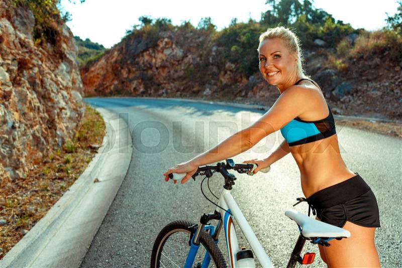 Happy woman cyclist standing near a bike on a mountain road, stock photo