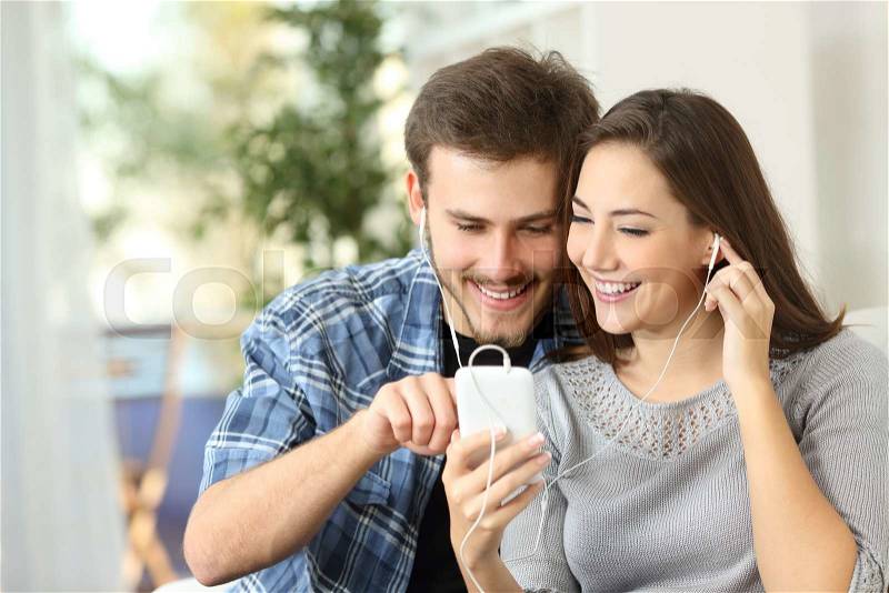 Happy couple sharing music from smart phone sitting on a couch at home, stock photo