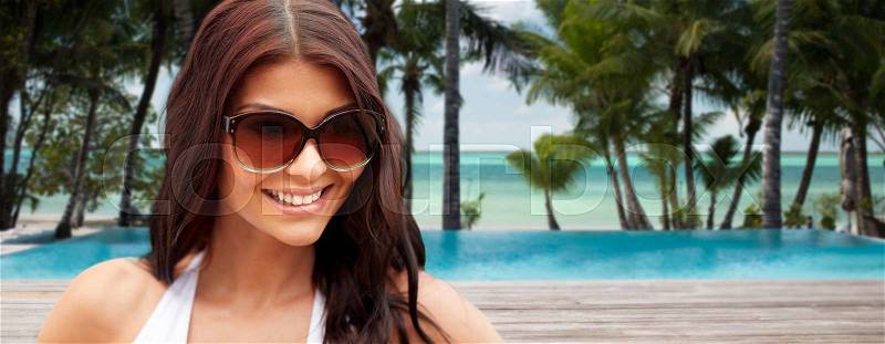 Summer vacation, tourism, travel, holidays and people concept -face of smiling young woman with sunglasses over tropical beach with palms and swimming pool background, stock photo