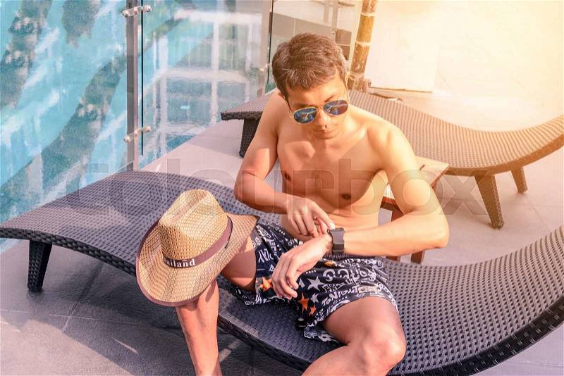Asian young man in sun sunglasses touching smart watch near swimming pool in summer, stock photo