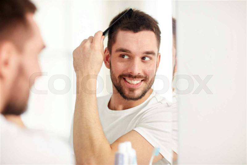 Beauty, grooming and people concept - smiling young man looking to mirror and brushing hair with comb at home bathroom, stock photo