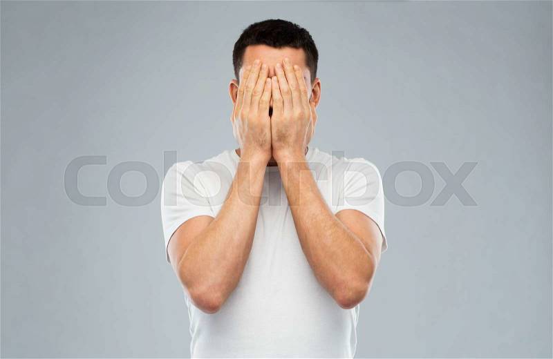 People, crisis, emotions and stress concept - man in white t-shirt covering his face with hands over gray background, stock photo