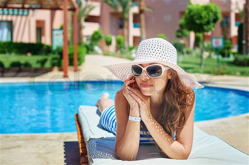 Beautiful woman in a big white hat on a lounger by the pool, stock photo