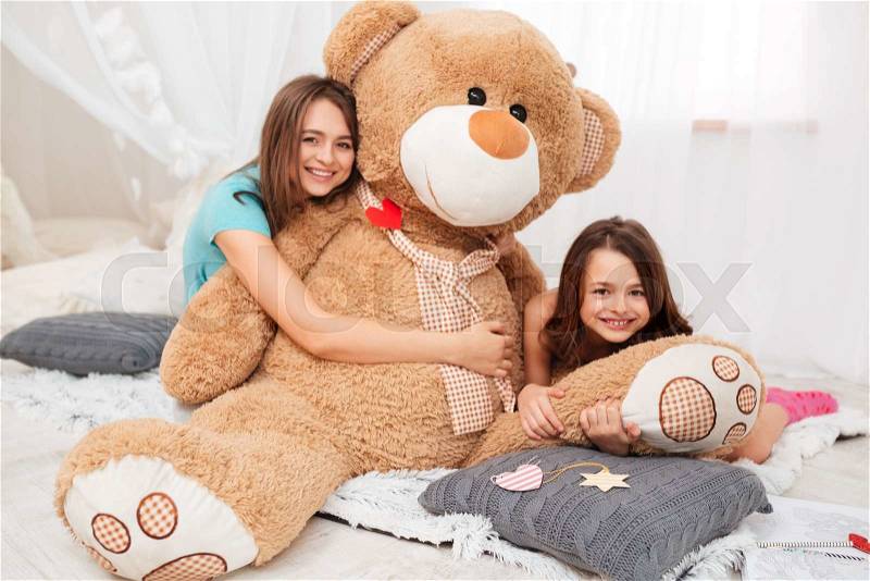 Two happy lovely sisters hugging plush bear in playroom, stock photo