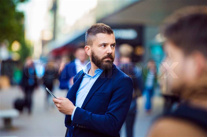 Hipster manager holding a smartphone outside in the crowded street, stock photo