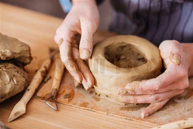 Hands of young woman making earthen pot on wooden table in pottery workshop, stock photo