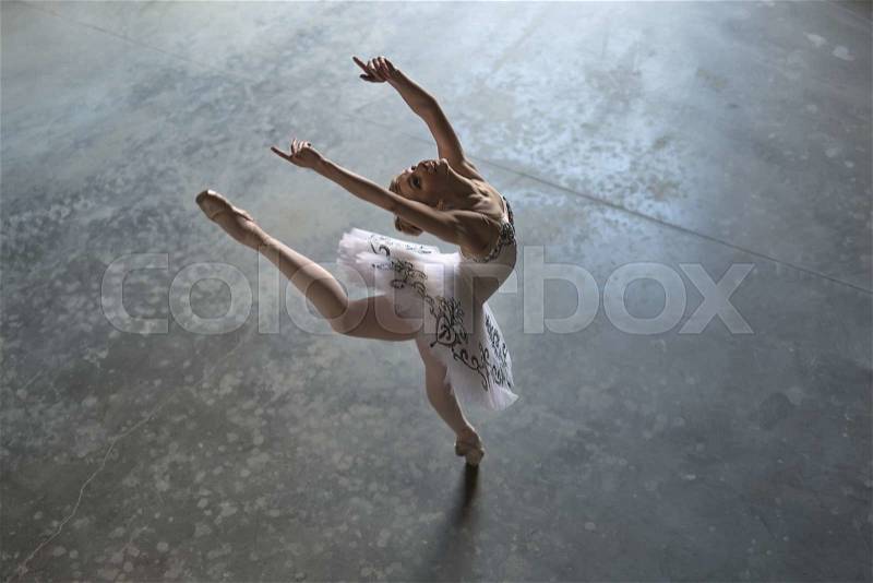 Pretty ballerina in a white tutu making a pose in a hall. She outstretches her hands and one leg up during tilting back. Daylight falls on her. Indoor. Horizontal, stock photo