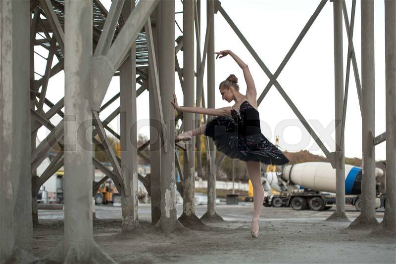 Nice ballerina posing leaning on a concrete support structure. She stands on the toe of the left foot in black tutu. Behind her is a parking for technical cars. Outdoor. Horizontal, stock photo