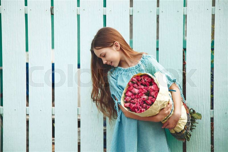 Tween girl holding a bunch of pink roses wrapped in craft paper over blue wooden fence, stock photo